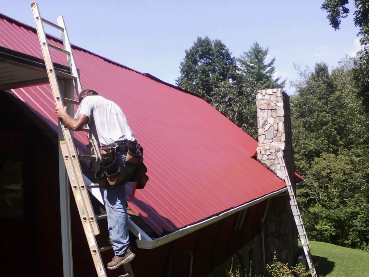 Roofer on a ladder beside a red metal roof and stone chimney — Soffit & Fascia Repair by Roofing Brothers in Milo